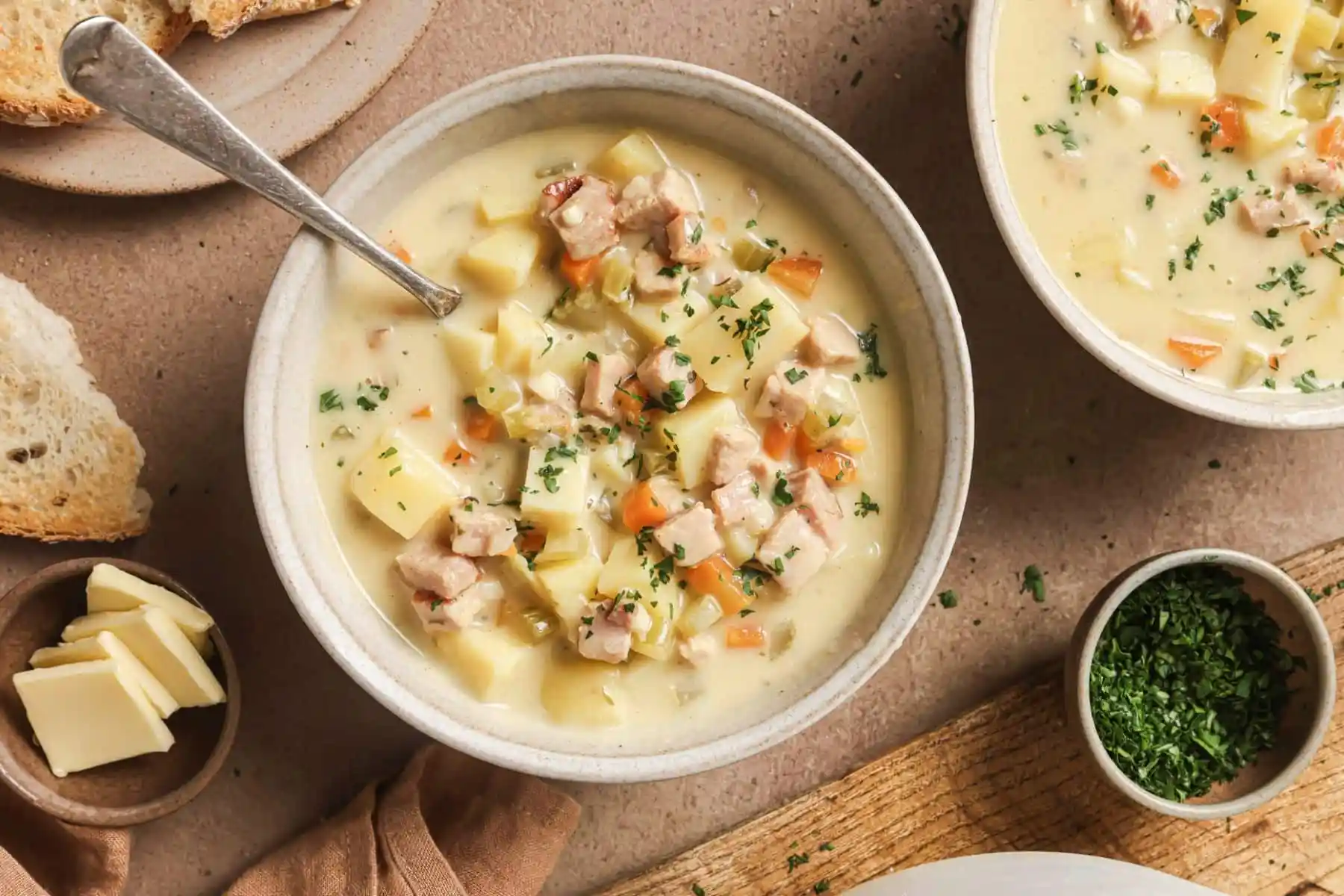A steaming bowl of creamy ham and potato soup with visible potato chunks, diced ham, and chopped parsley on top, set on a rustic wooden table with a spoon nearby