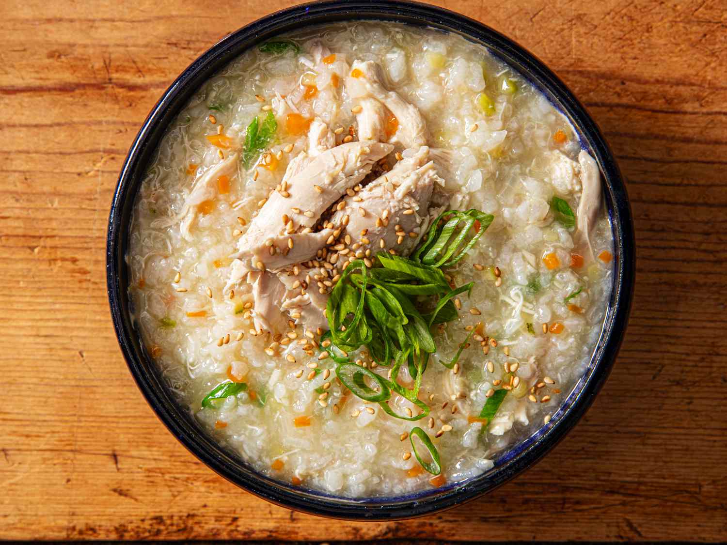 A steaming bowl of creamy rice porridge on a wooden table, topped with sliced scallions, sesame oil, and a soft egg, with a spoon beside it in warm natural light