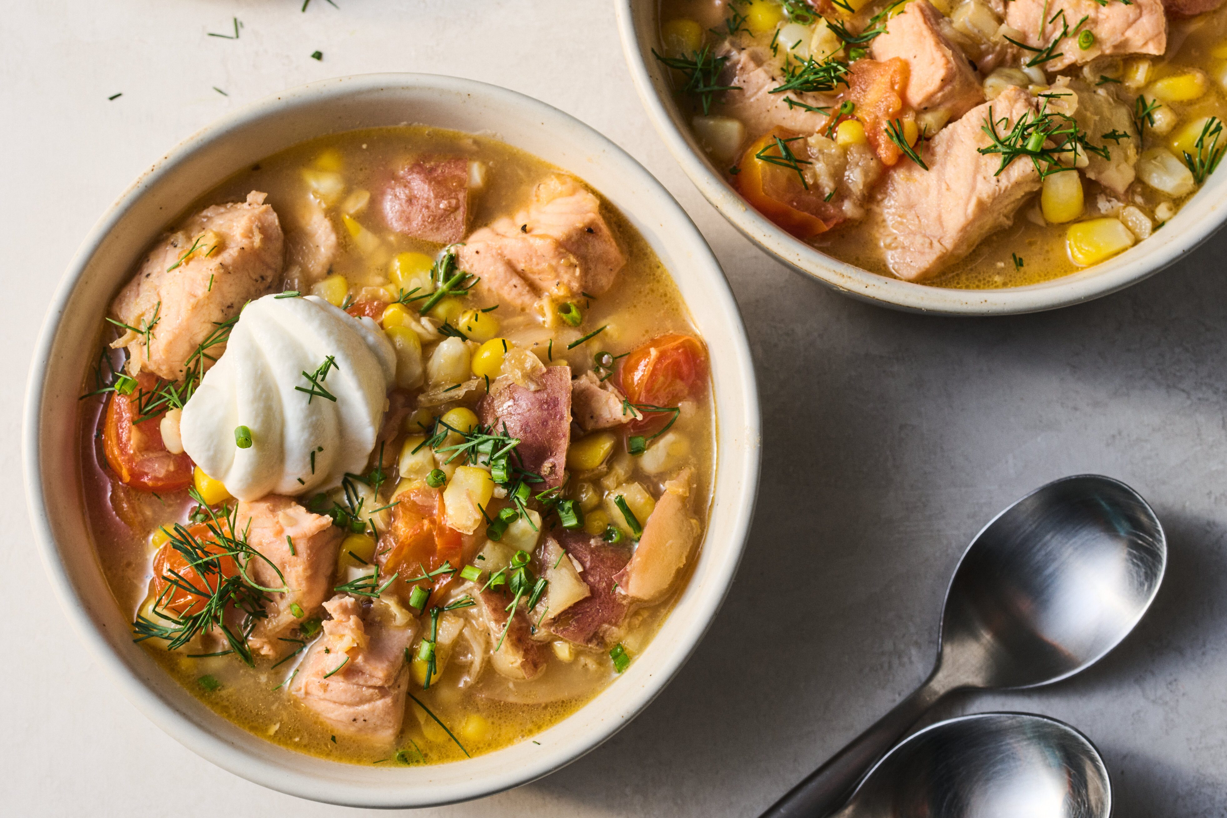 A steaming bowl of creamy salmon chowder with chunks of salmon, potatoes, and corn, topped with fresh dill and cracked black pepper on a wooden table with a spoon beside it