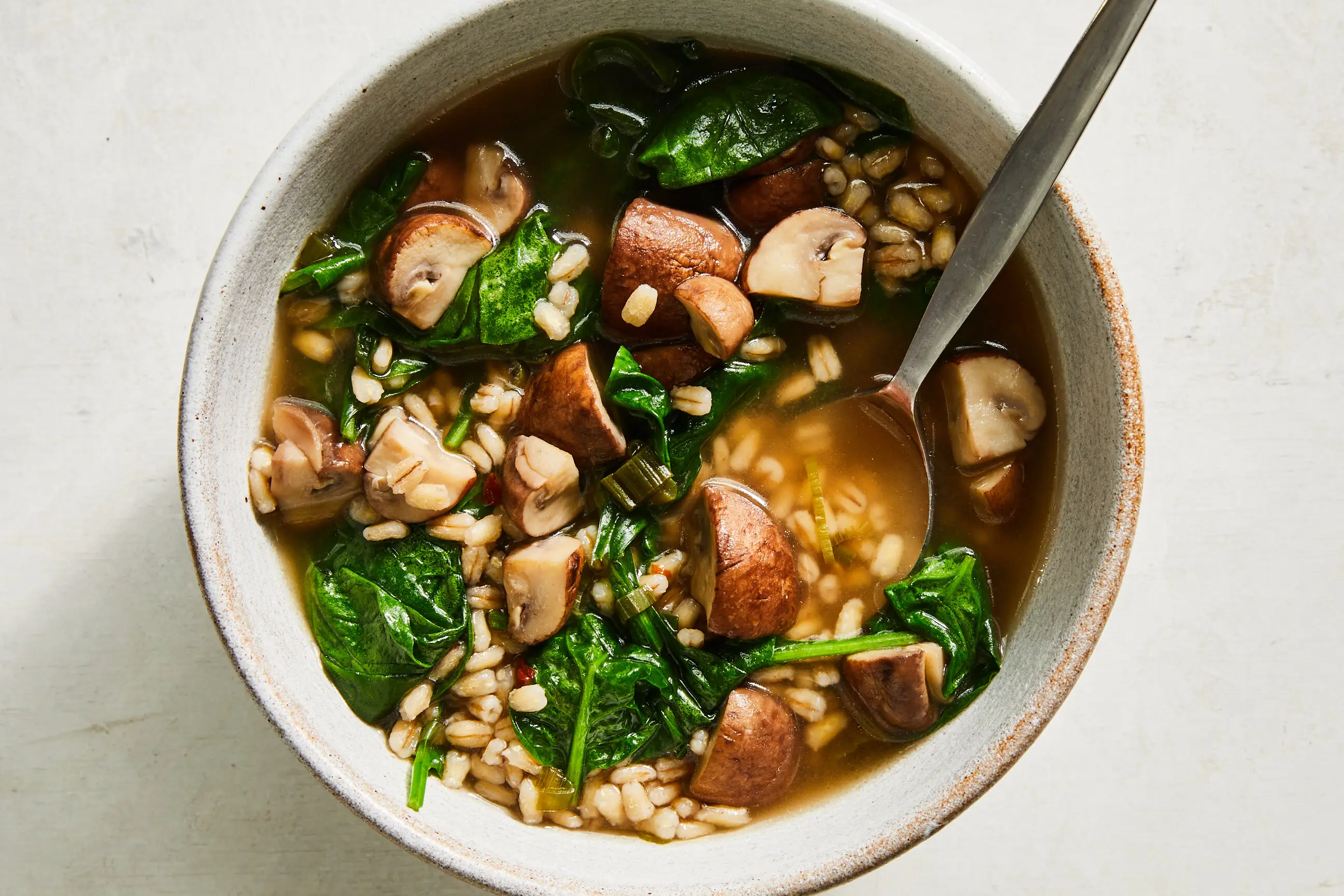 A steaming bowl of mushroom barley soup with visible sliced mushrooms and barley grains, topped with chopped parsley on a rustic wooden table, warm kitchen lighting