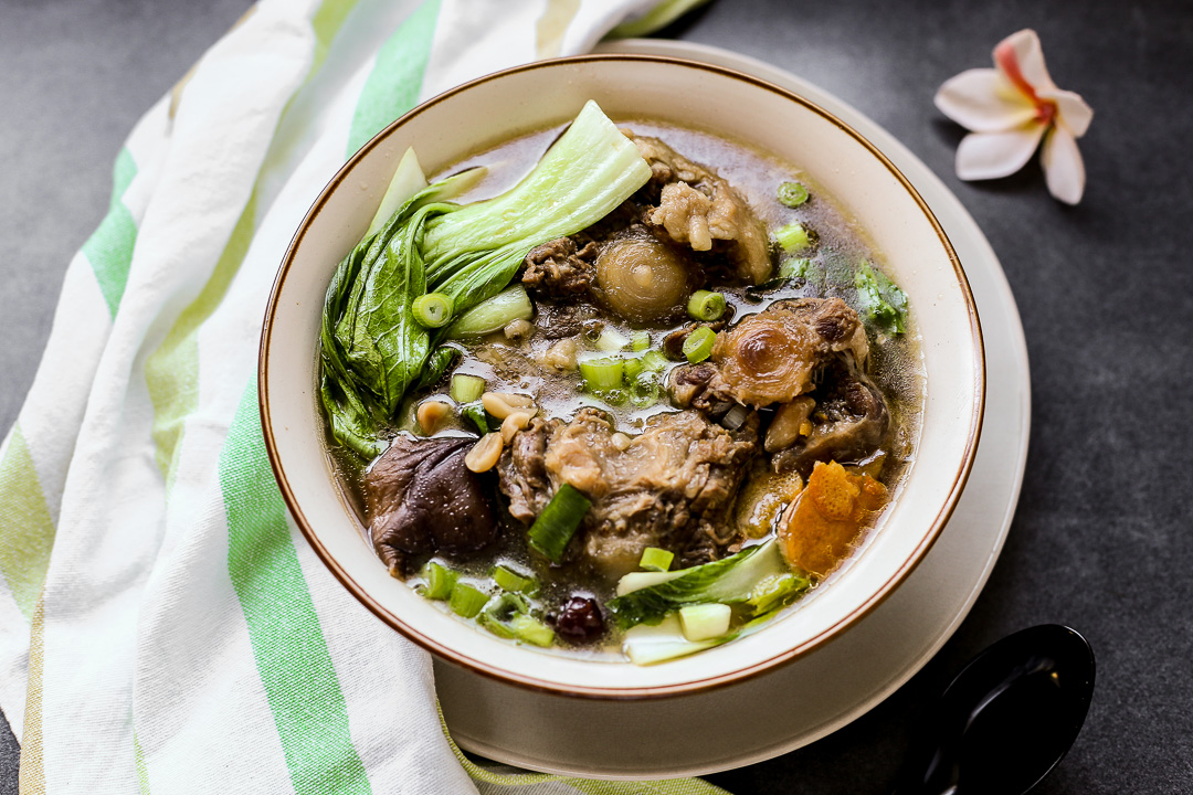 A steaming bowl of oxtail soup with tender oxtail pieces, carrots, celery, and fresh parsley on top, set on a wooden table with a spoon and napkin