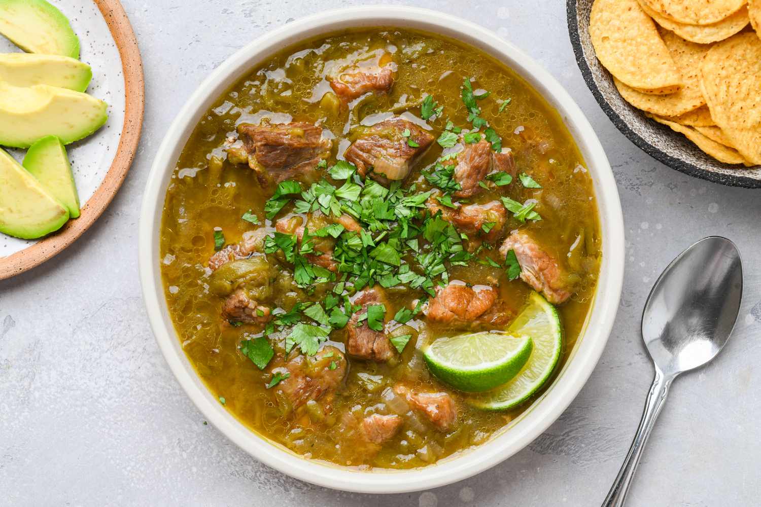 A steaming bowl of pork green chili with roasted green chile pieces, tender pork chunks, and a sprinkle of chopped cilantro on top, set on a wooden table with warm natural kitchen light