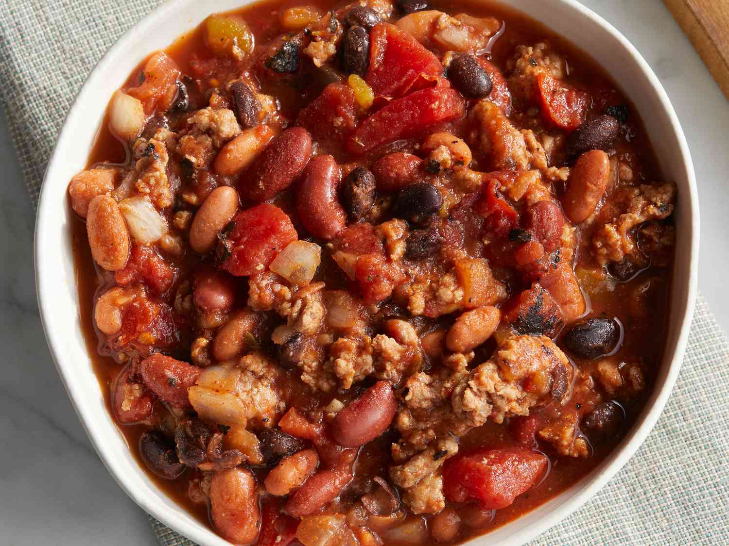 A steaming bowl of smoky kidney bean chili topped with diced red onion and cilantro on a rustic wooden table with a lime wedge