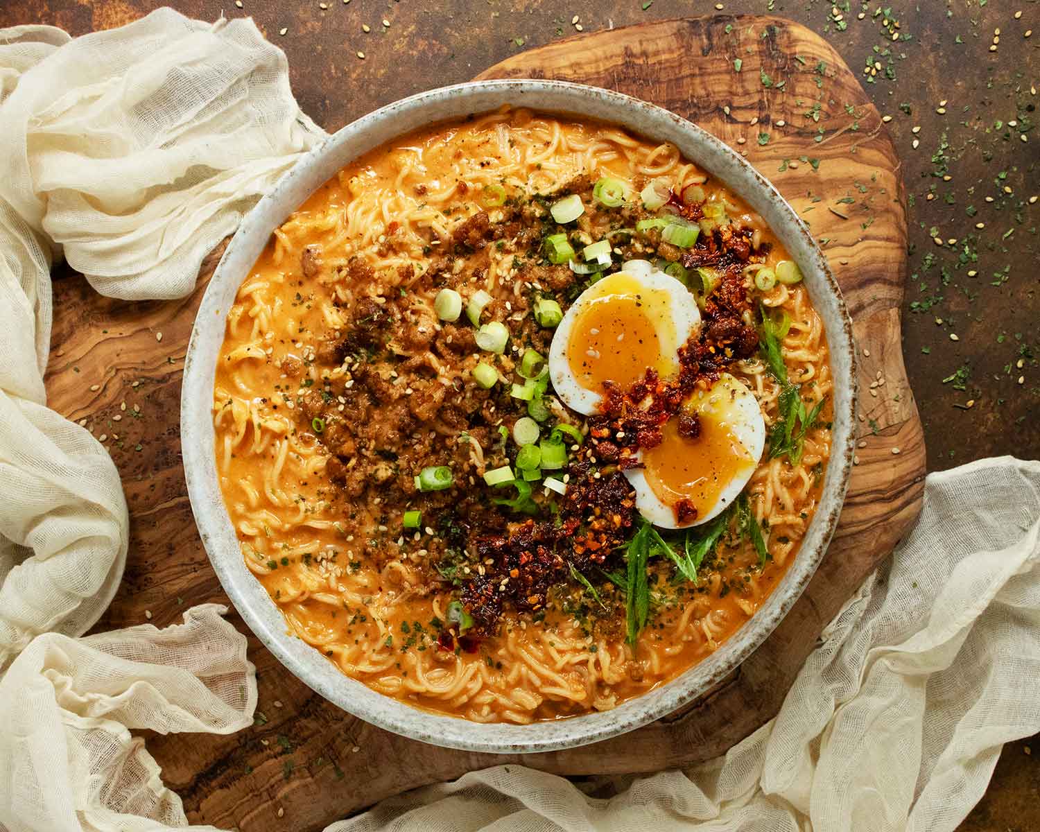 A steaming bowl of spicy garlic peanut ramen topped with sliced scallions, sesame seeds, and a jammy egg on a simple wooden kitchen table with soft natural light