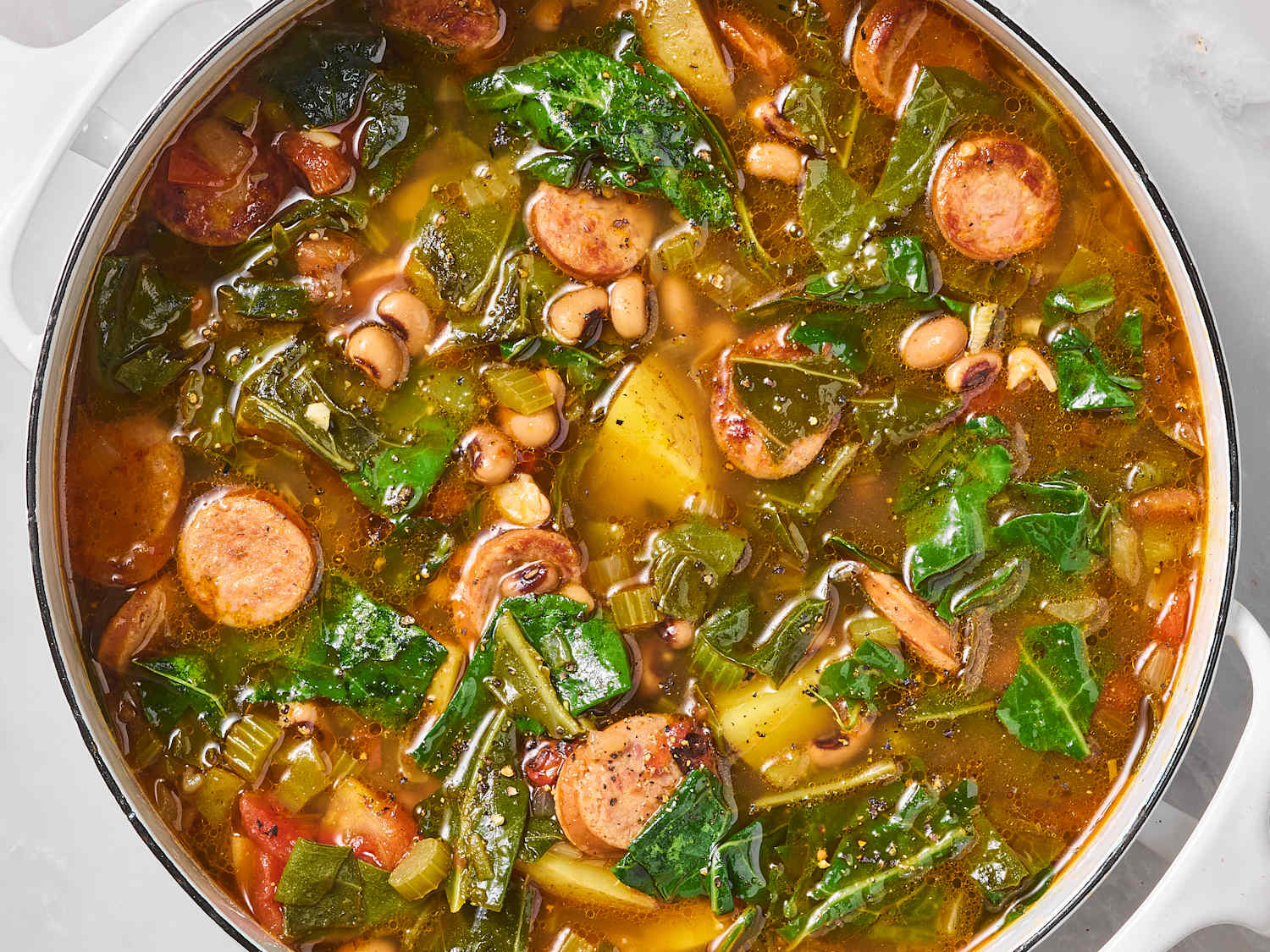 A steaming bowl of swamp soup with sliced smoked sausage, white beans, and dark leafy greens in a rustic ceramic bowl on a wooden table with a spoon beside it