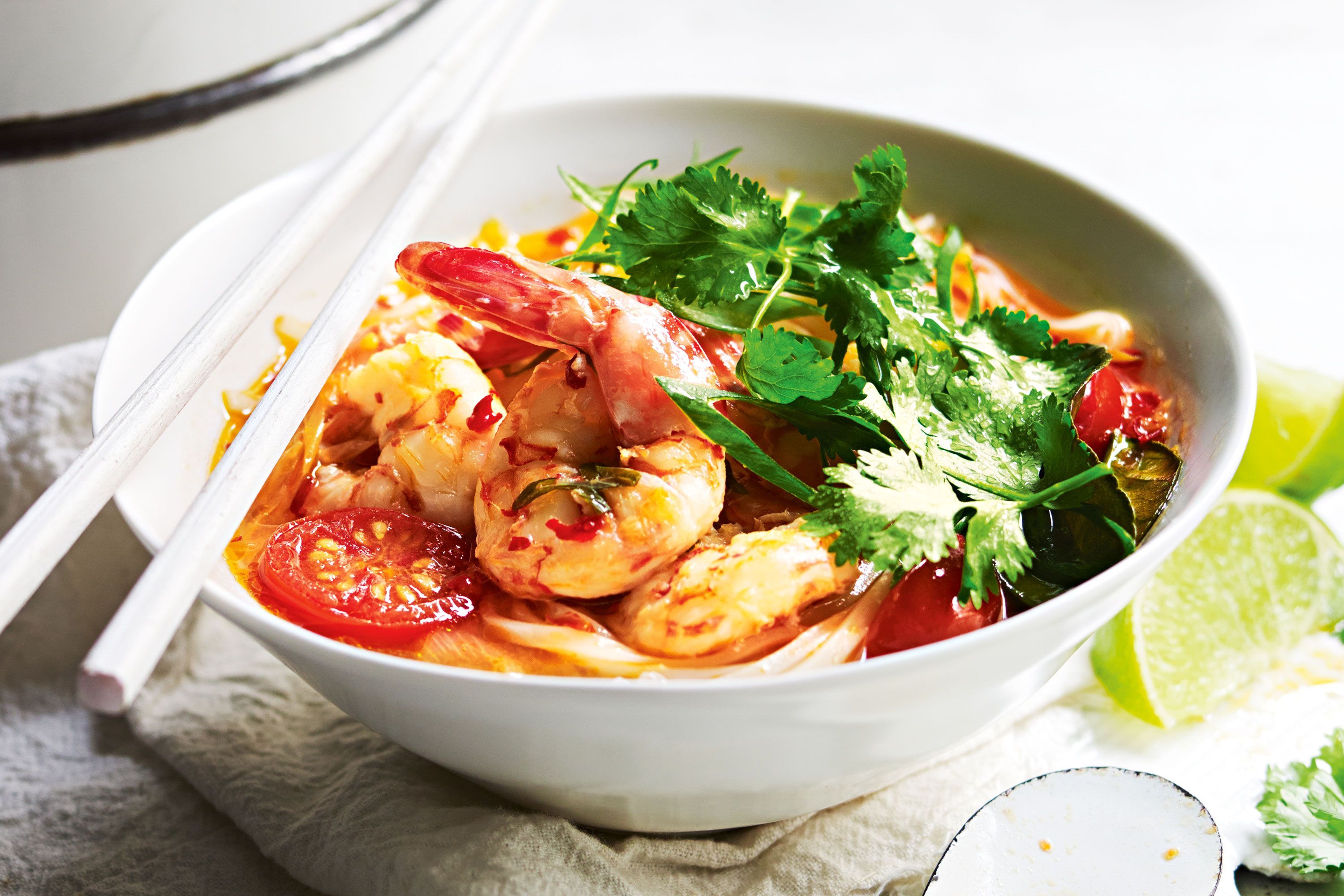A steaming bowl of tom yum soup with shrimp, mushrooms, cilantro, and red chili slices on a rustic table with lime wedges, natural kitchen lighting