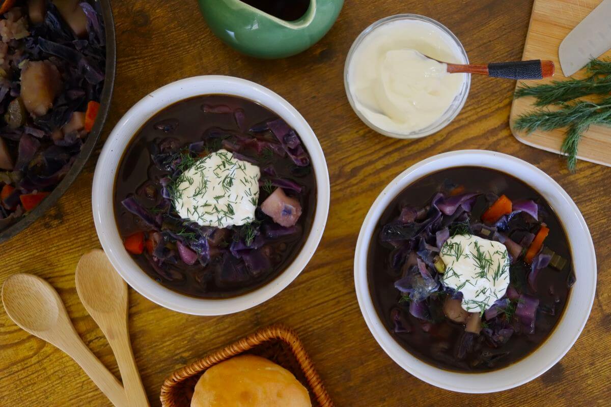 A steaming bowl of tomato-based cabbage soup with carrots, celery, and cabbage in a white bowl on a wooden table, spoon nearby, natural kitchen light