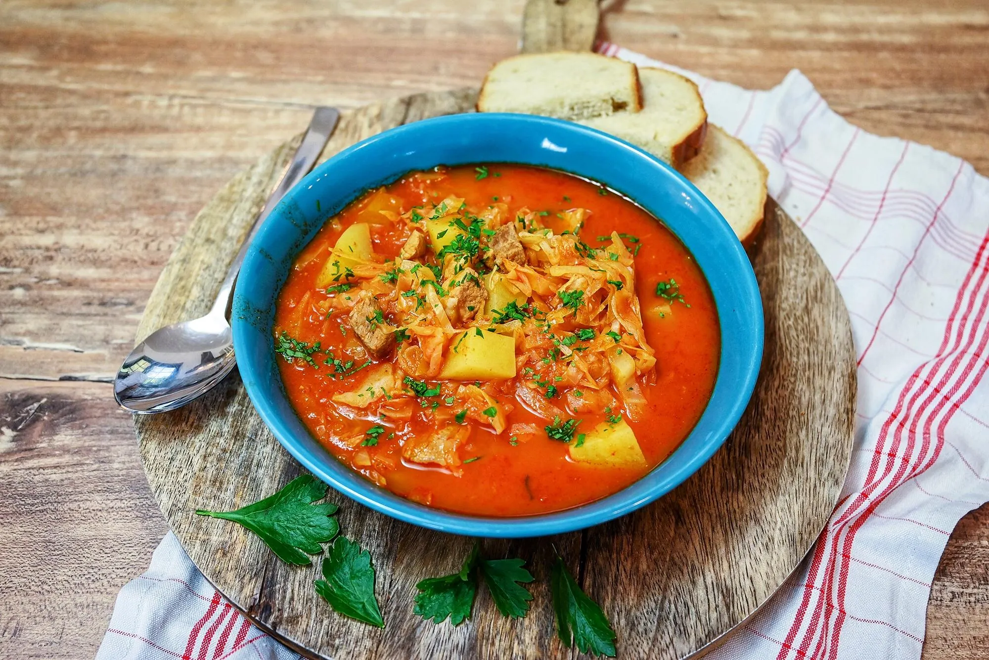 A steaming bowl of tomato-based cabbage soup with visible cabbage ribbons, carrots, celery, and herbs on a wooden table with a spoon nearby, warm natural kitchen lighting