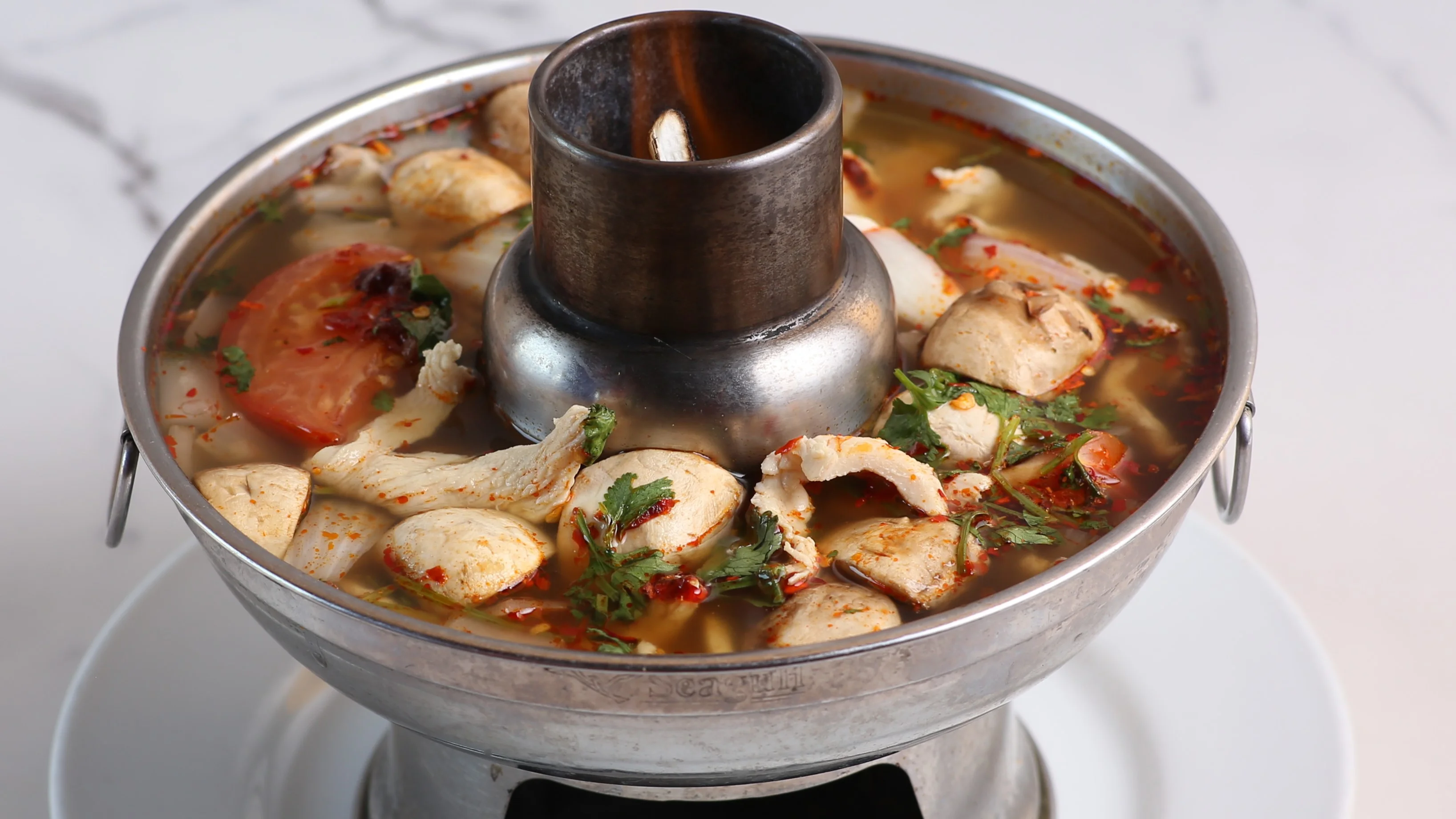 A steaming bowl of tomyam soup with shrimp, mushrooms, cilantro, and red chili slices on a wooden table with a spoon and lime wedge