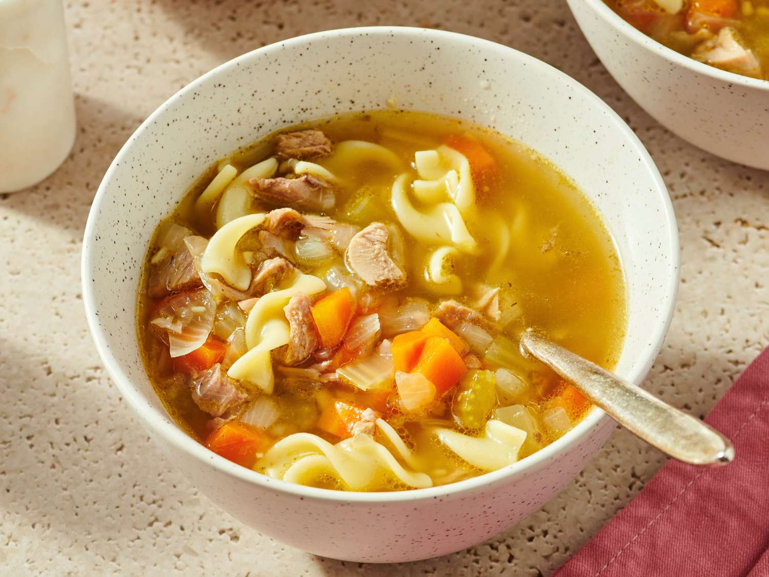 A steaming bowl of turkey noodle soup with egg noodles, shredded turkey, carrots, celery, and fresh parsley on a light stoneware table