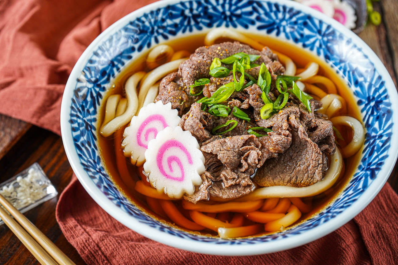 A steaming bowl of udon noodle soup with mushrooms, bok choy, sliced scallions, and a soft-boiled egg in a neutral stoneware bowl on a warm kitchen table