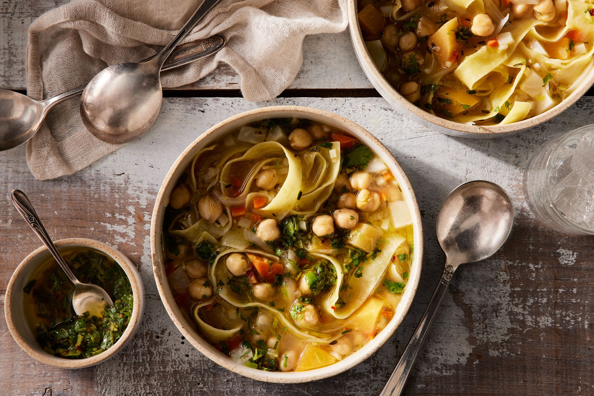 A steaming bowl of vegan chickpea noodle soup with carrots, celery, noodles, fresh dill, and a lemon wedge on a stone-colored kitchen counter in warm natural light