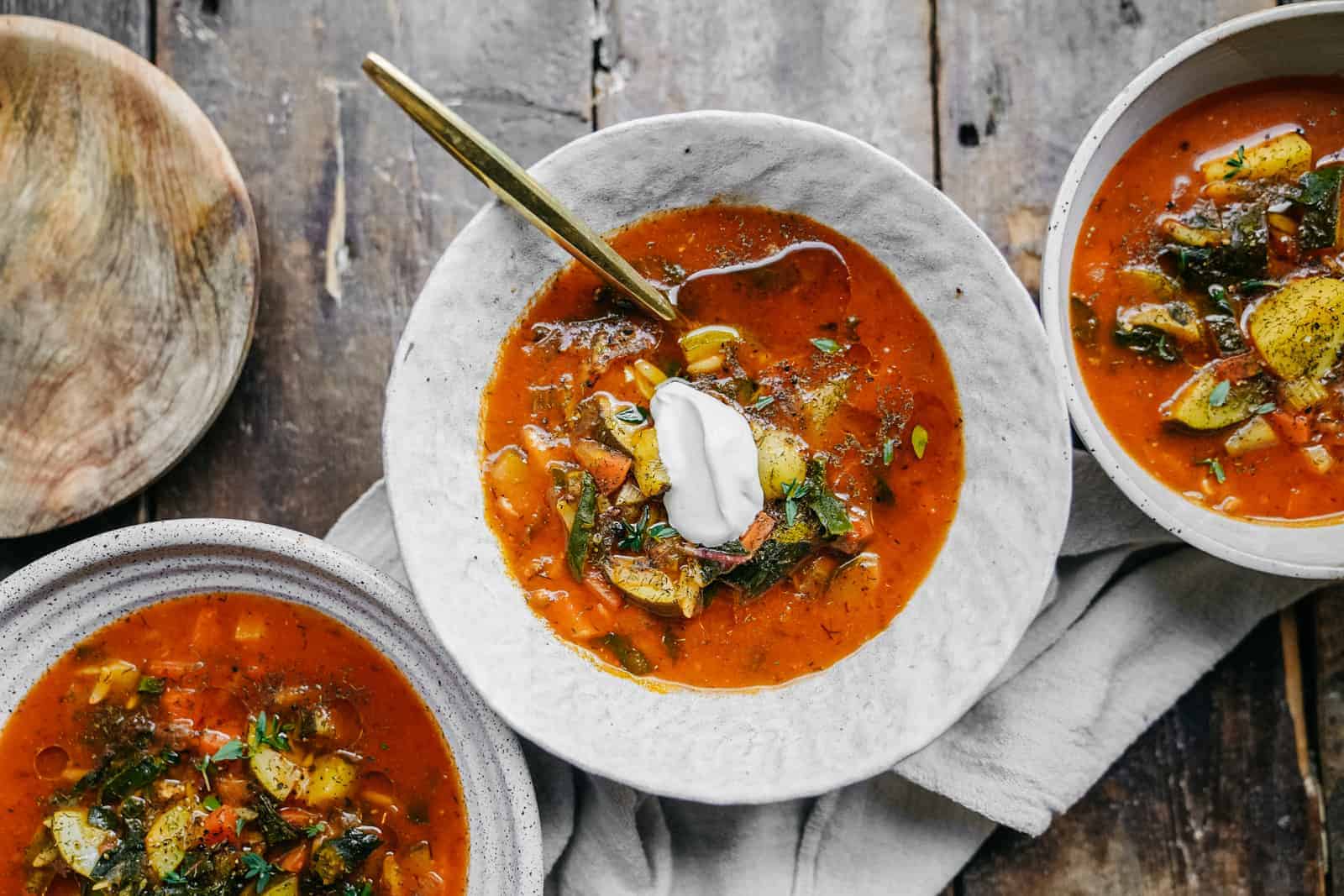 A steaming bowl of vegetable soup topped with fresh parsley on a stoneware table with a spoon and a linen napkin, warm kitchen lighting