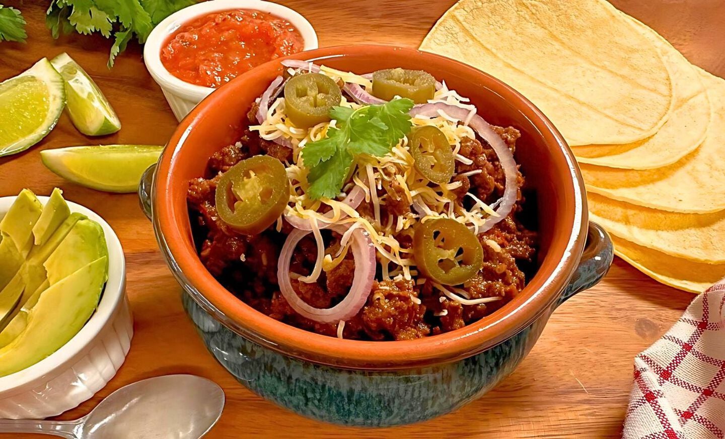A steaming bowl of venison chili topped with diced onion, shredded cheddar, and cilantro on a wooden kitchen table with warm natural light