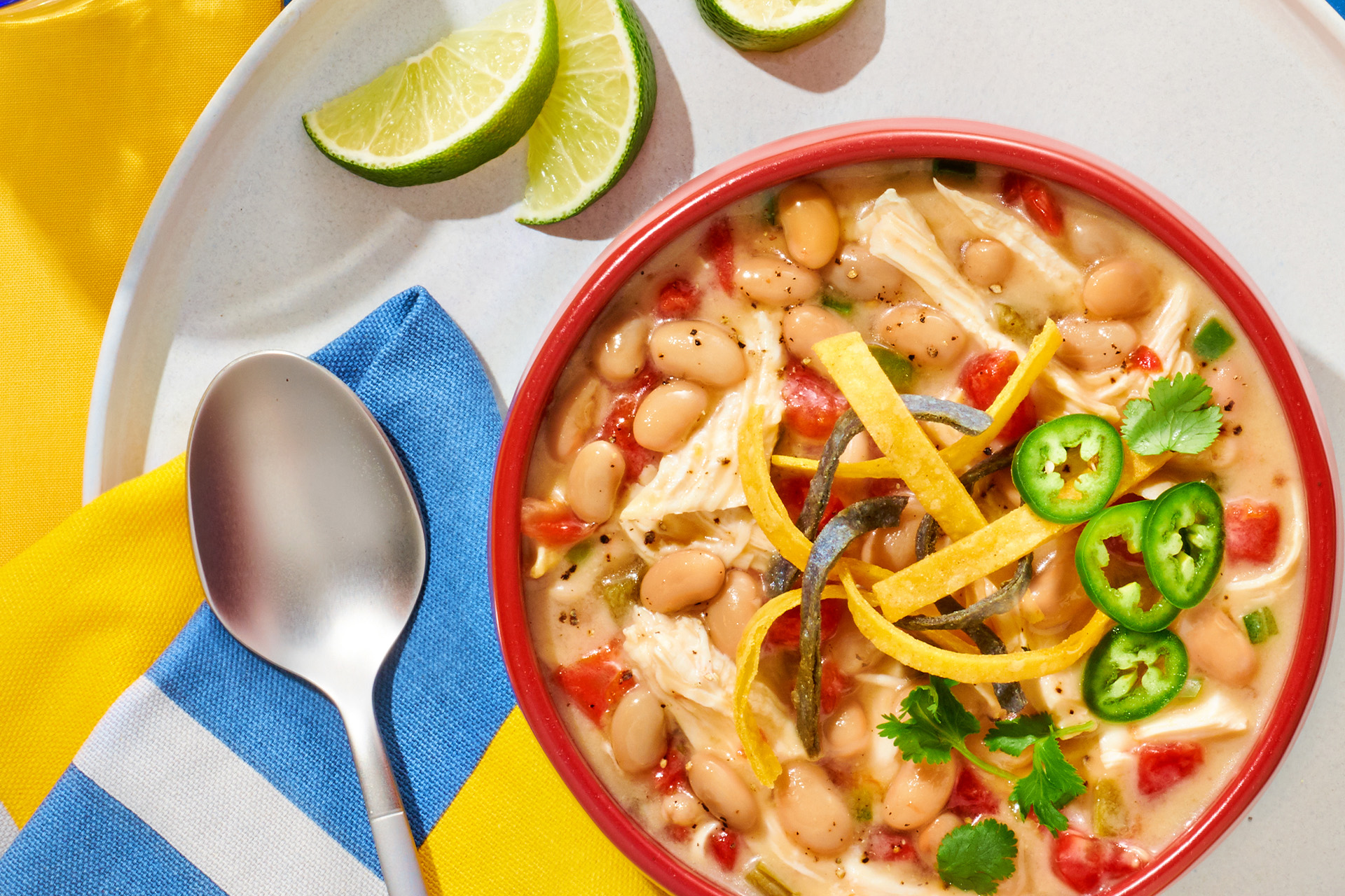 A steaming bowl of white bean and chicken chili topped with cilantro, sliced jalapeno, and a dollop of sour cream on a wooden table with lime wedges