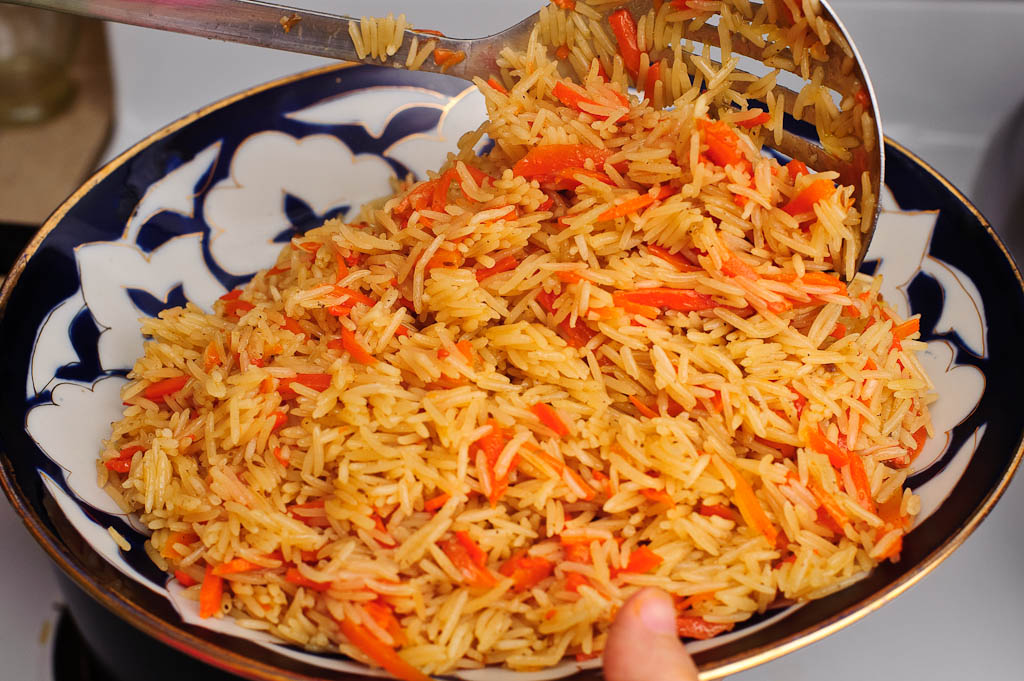 A steaming platter of chicken plov with fluffy rice, golden carrots, and a whole garlic head nestled on top, served on a rustic table with warm natural light