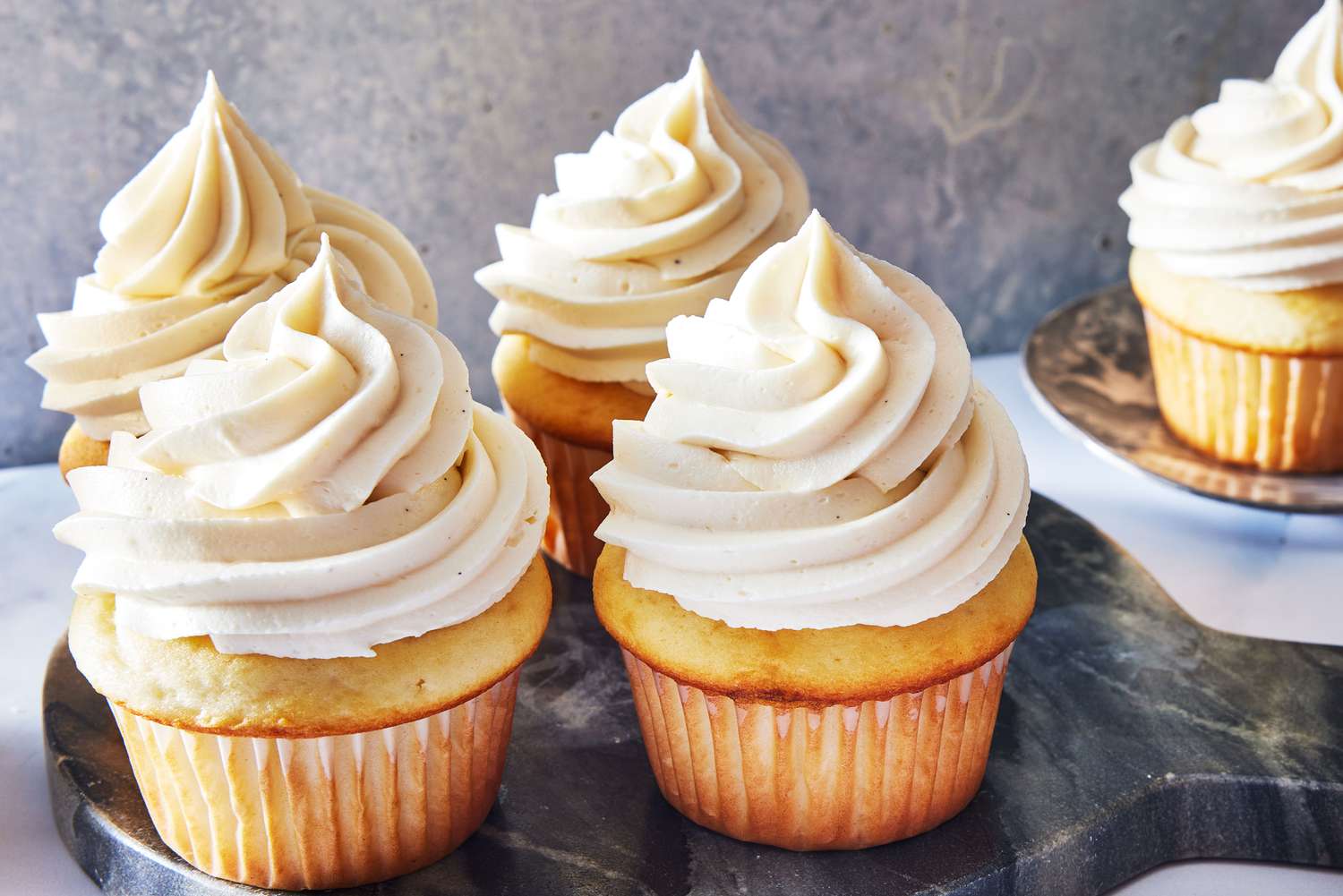 A swirl of pale vanilla buttercream piped high on a vanilla cupcake on a light kitchen counter with soft natural light