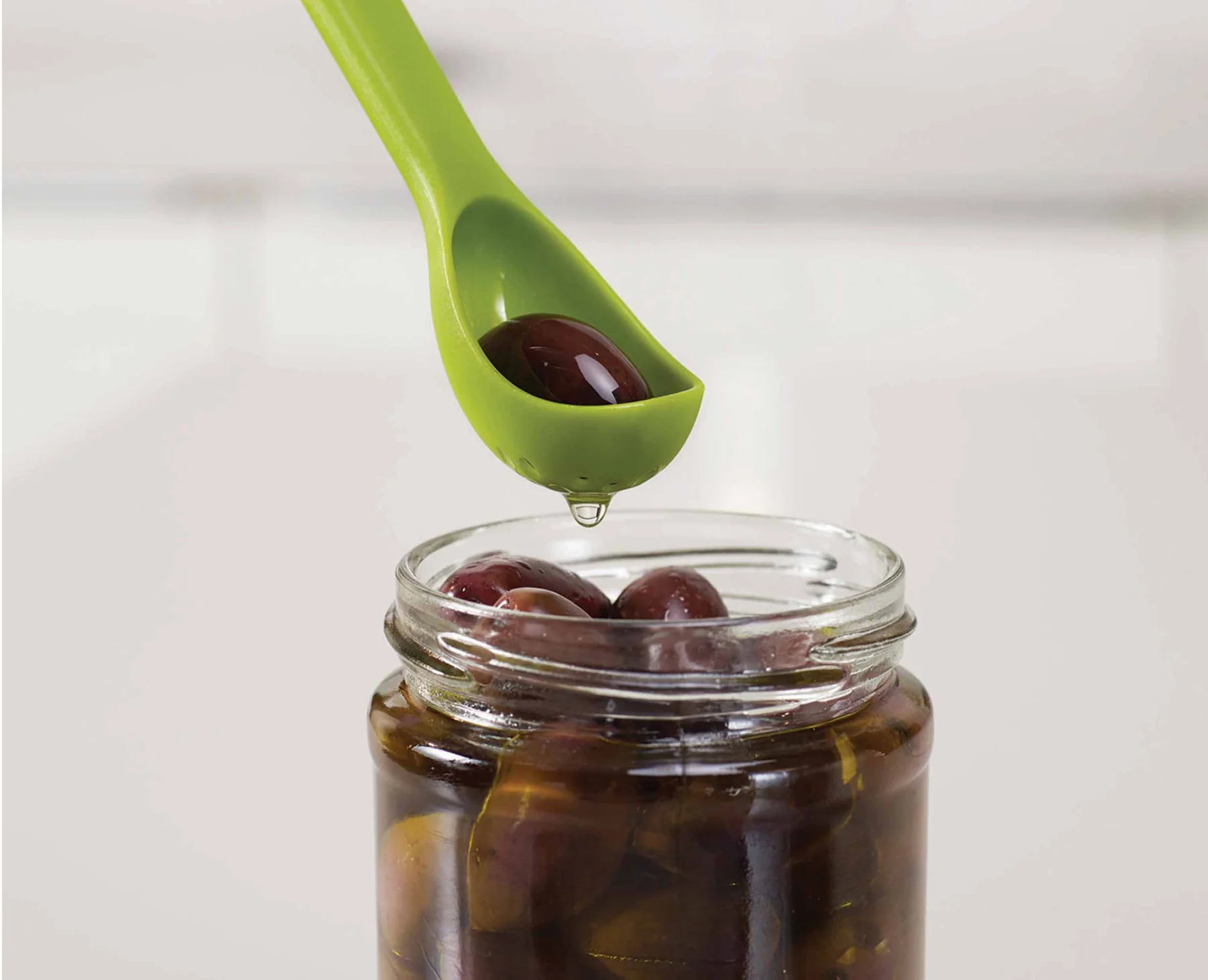 A tablespoon measuring spoon filled with pale green olive brine held over a cocktail shaker on a kitchen counter
