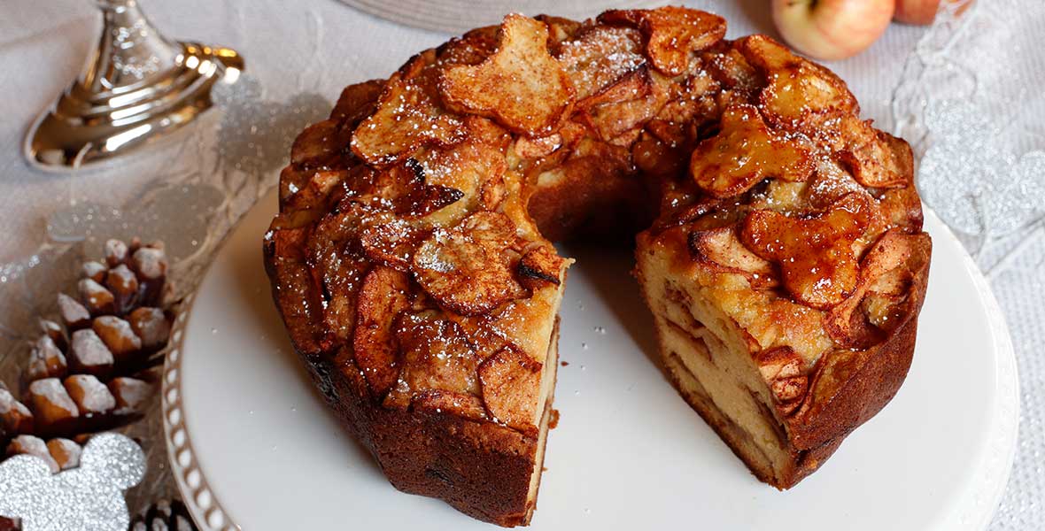 A tall Jewish apple cake on a simple cake stand, golden brown with a crackly sugar top, sliced to show cinnamon apple layers, set on a warm-toned kitchen table with soft window light