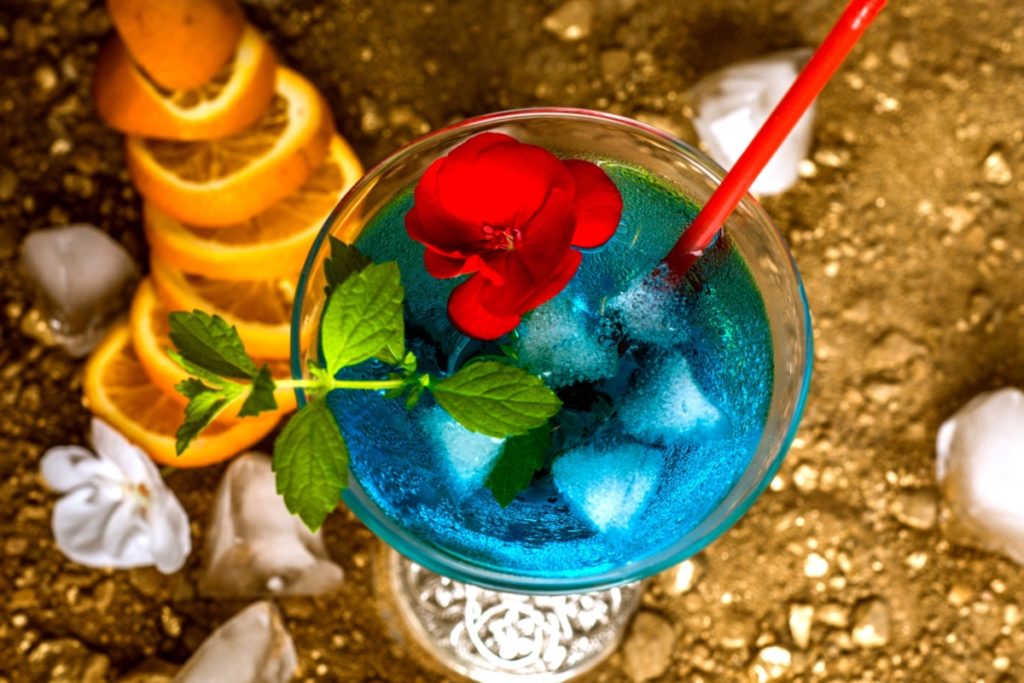 A tall blue AMF cocktail in a highball glass as a spoon stirs through ice, with condensation on the glass on a kitchen counter