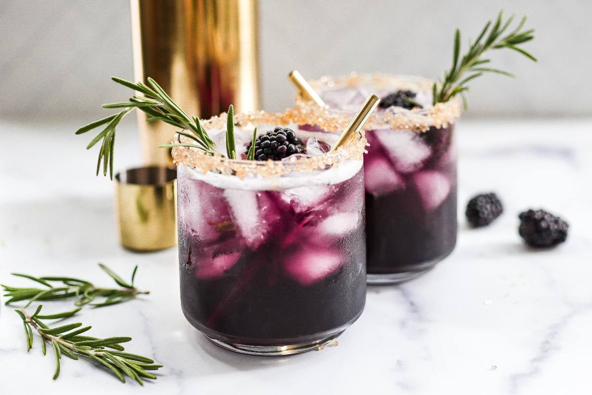 A tall clear glass filled with a deep purple berry mocktail over ice, topped with sparkling bubbles and a subtle creamy swirl, on a warm kitchen counter with soft natural light