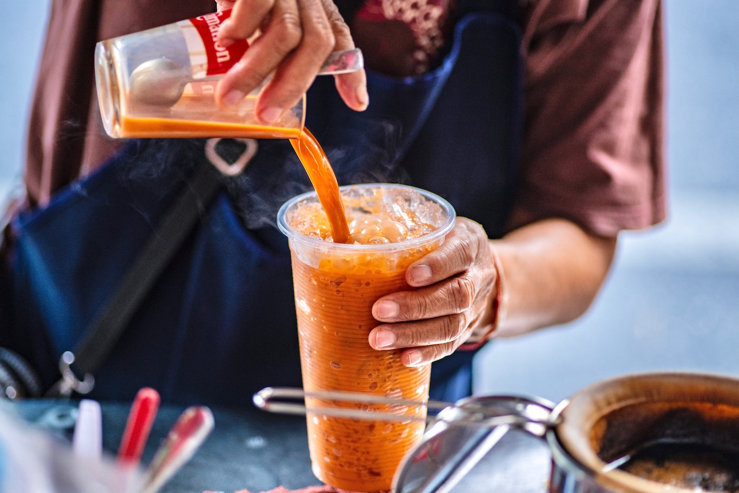 A tall clear glass of Thai iced tea with lots of ice, a creamy layer on top, and condensation on the outside, set on a light kitchen counter with warm afternoon light