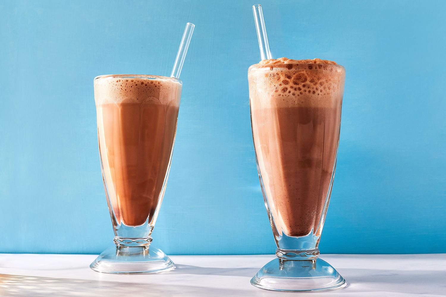 A tall clear glass on a kitchen counter with a long spoon nearby, ready for making an egg cream
