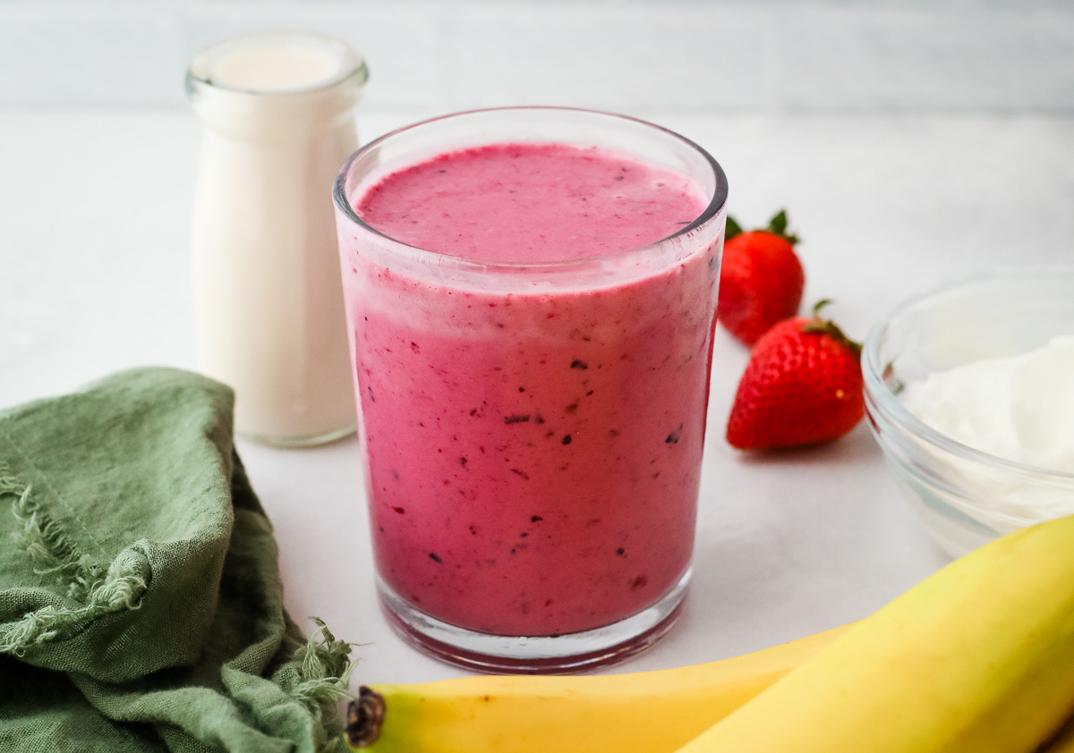 A tall glass of creamy banana berry smoothie on a bright kitchen counter with a metal straw and a few sliced bananas nearby