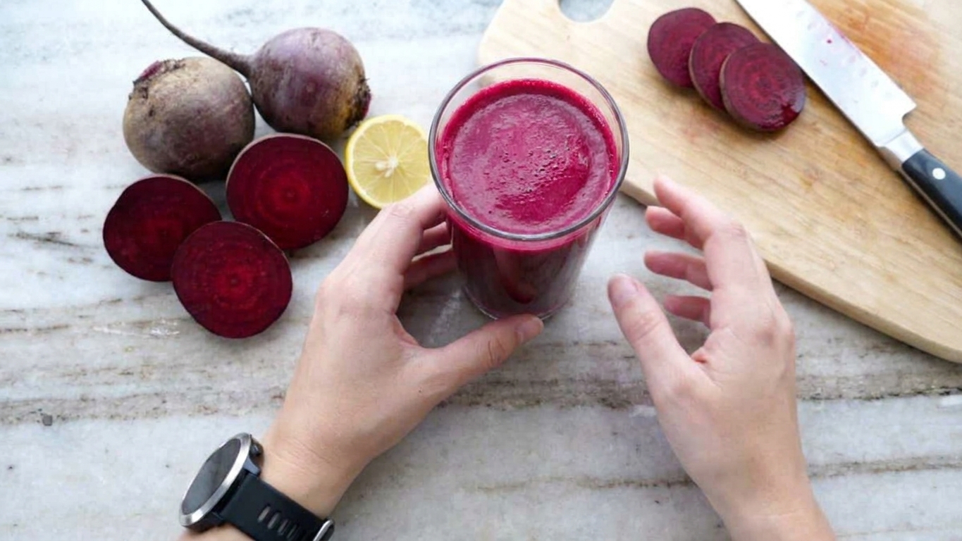 A tall glass of deep magenta beetroot juice on a light counter with whole beets and a lemon nearby