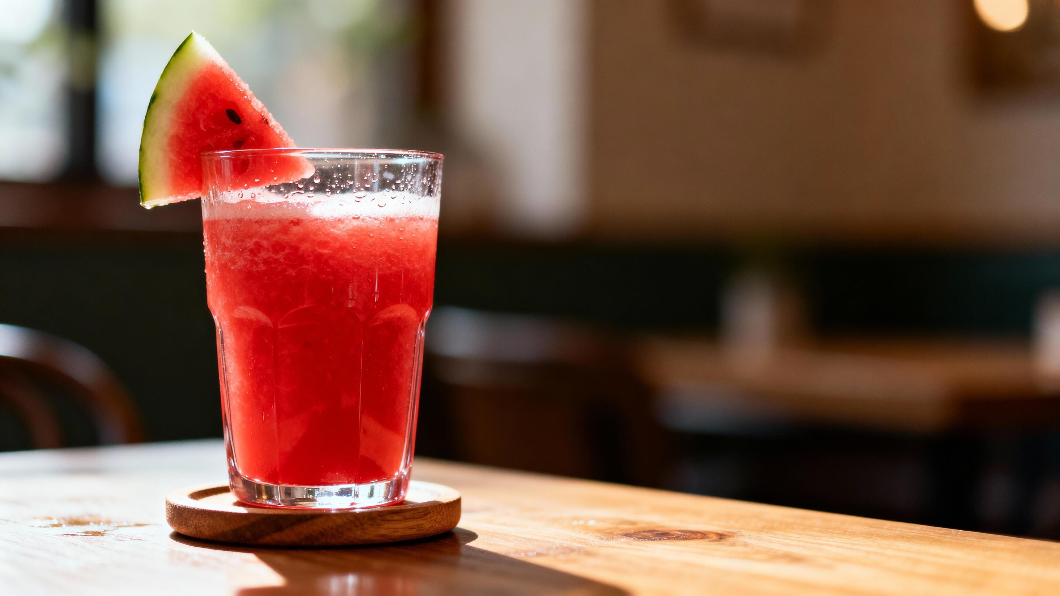 A tall glass of fresh watermelon juice with condensation on the outside, set on a light stone kitchen counter with a few watermelon wedges nearby