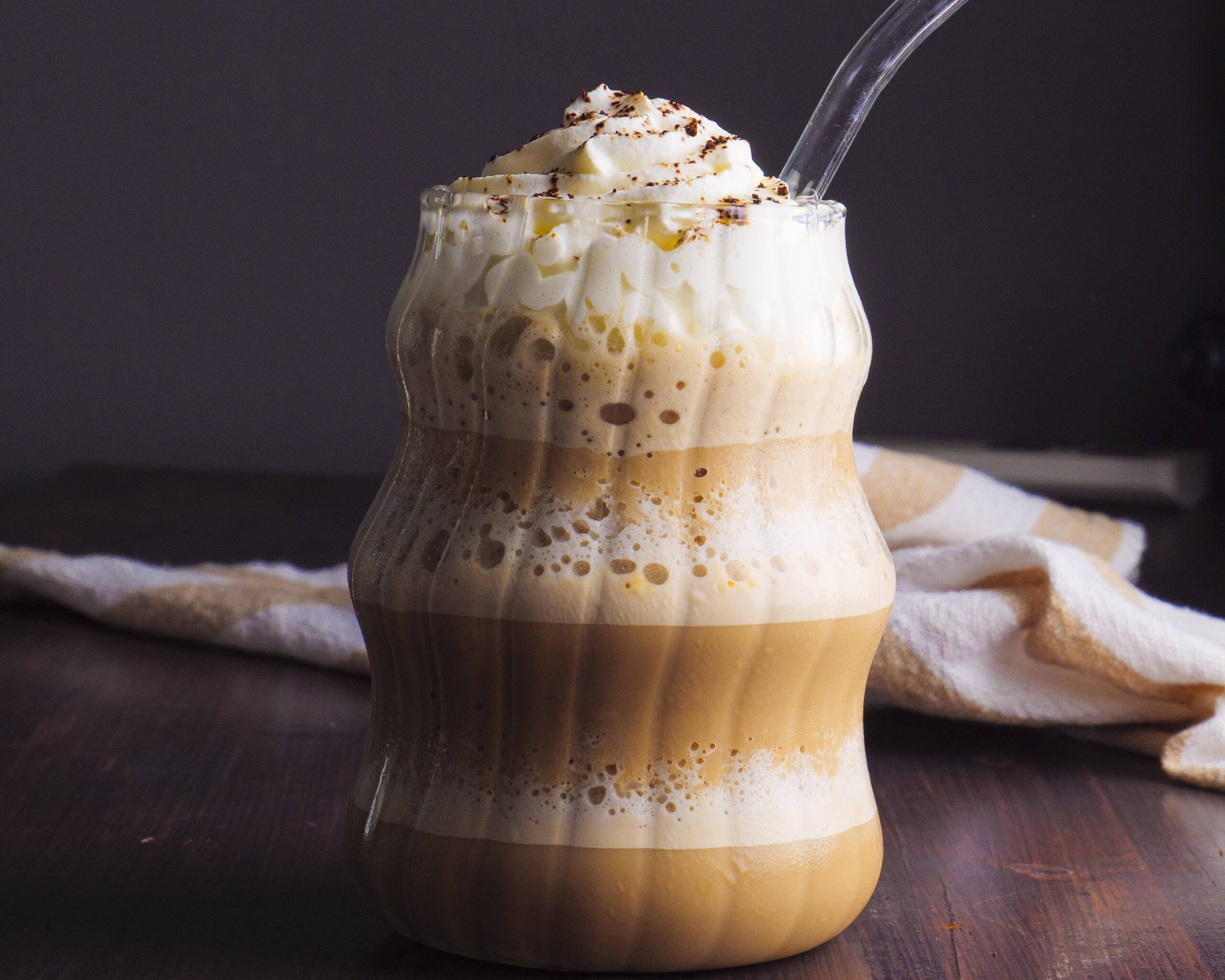 A tall glass of homemade coffee Frappuccino topped with whipped cream and a light drizzle of chocolate syrup on a bright kitchen counter