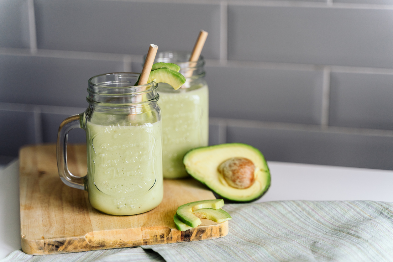 A tall glass of pale green avocado smoothie on a light stone kitchen counter with a lime wedge and a few ice cubes nearby, soft natural window light