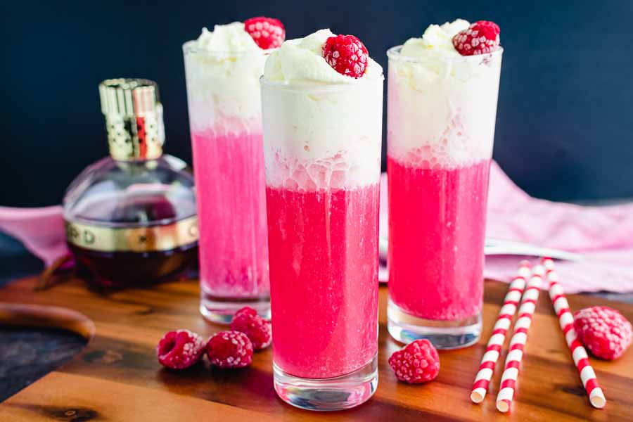 A tall glass of raspberry Italian soda with ice, sparkling bubbles, and a light cream swirl on a sunlit kitchen counter