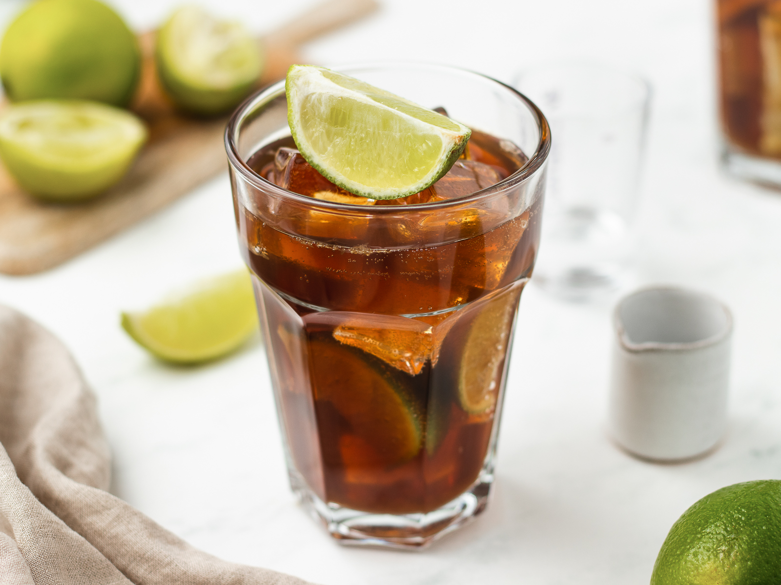 A tall glass of rum and Coke packed with clear ice cubes, a lime wedge on the rim, and condensation on the glass on a clean kitchen counter