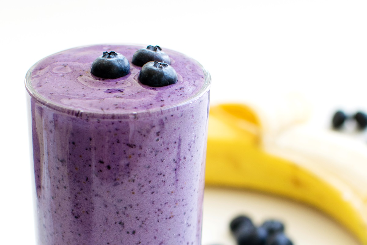 A tall glass of thick blueberry smoothie on a light kitchen counter with a few fresh blueberries scattered nearby and soft morning light