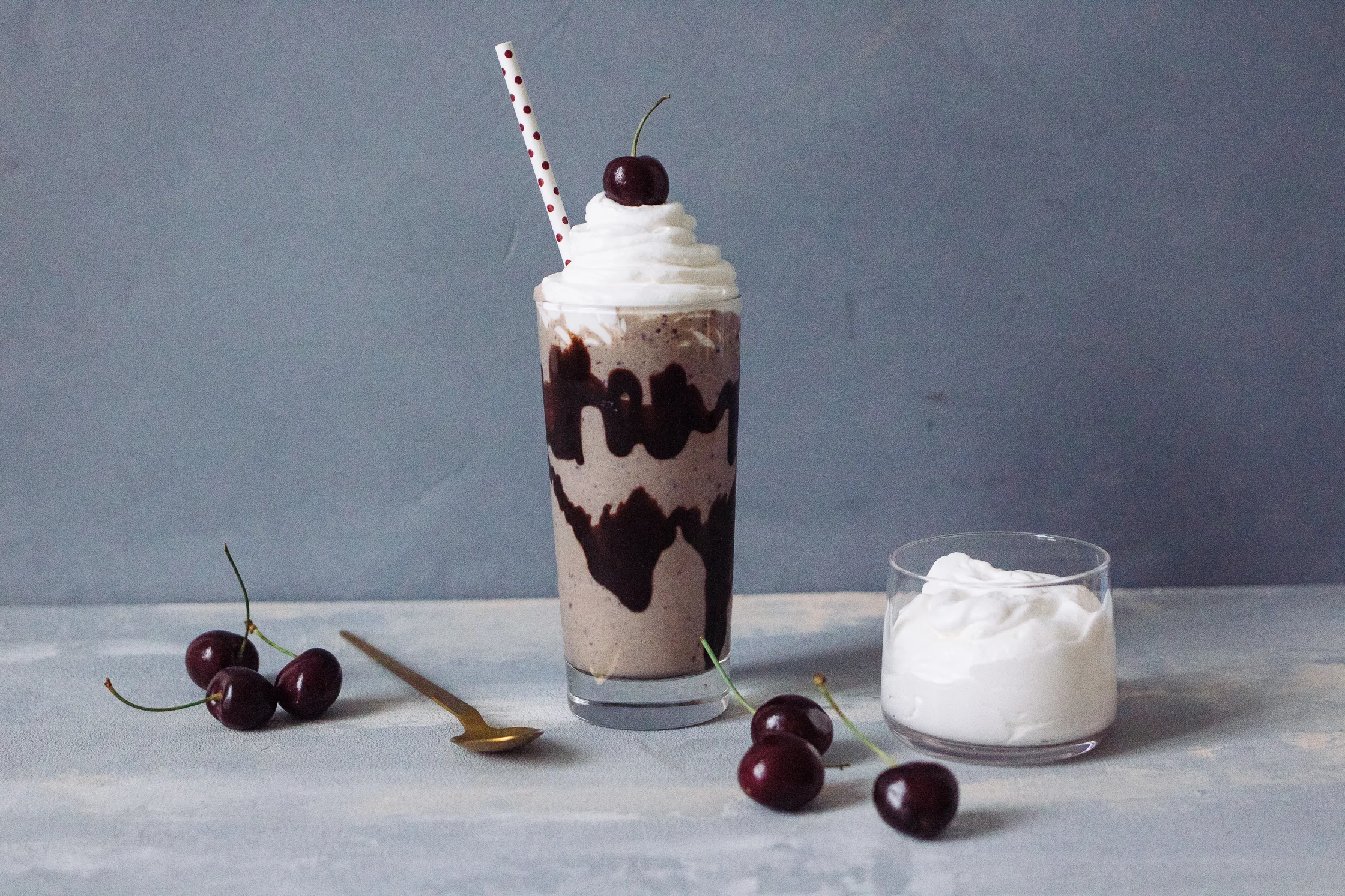 A tall glass of thick vanilla milkshake topped with whipped cream and a cherry on a wooden kitchen counter with a straw beside it