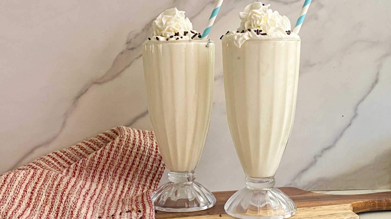 A tall glass of vanilla slushie milkshake with a frosty texture on the surface, whipped cream on top, and a striped straw on a neutral kitchen counter with soft natural light
