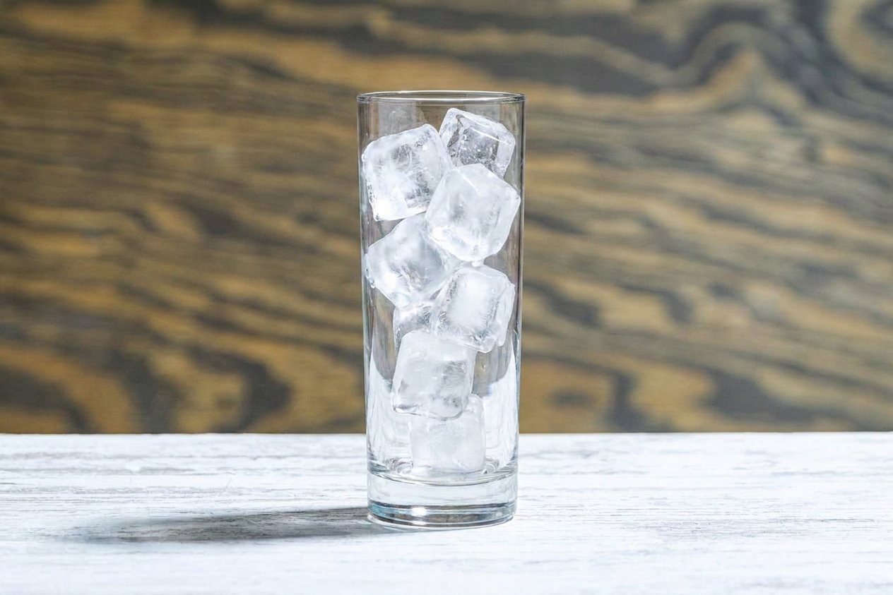 A tall highball glass filled with clear ice cubes on a wooden kitchen counter