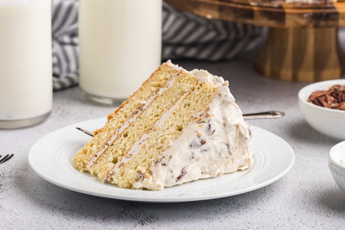 A tall slice of Italian cream cake on a white dessert plate, showing three coconut-pecan cake layers with cream cheese frosting, set on a warm kitchen table with soft natural light