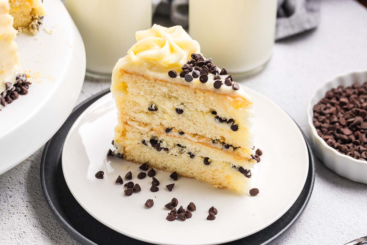 A tall slice of cannoli cake on a dessert plate, showing vanilla cake layers with ricotta mascarpone filling studded with mini chocolate chips, topped with a light dusting of powdered sugar on a bright kitchen table