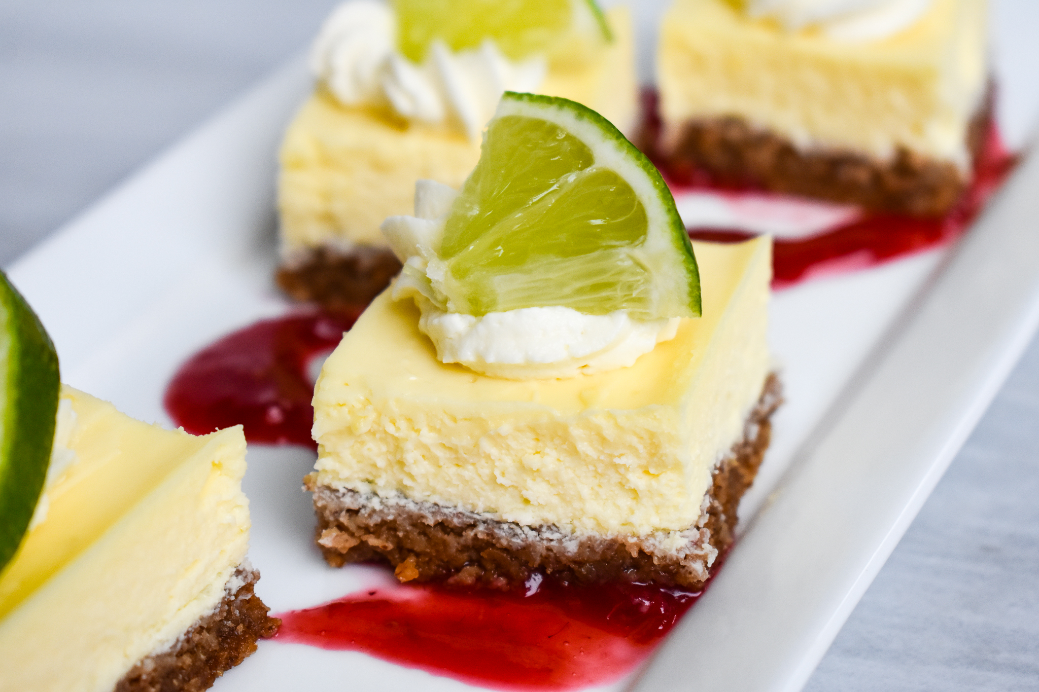 A tall slice of key lime cheesecake on a white plate with a graham cracker crust and a few lime zest curls on top