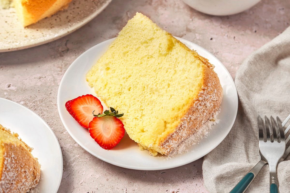 A tall slice of vanilla chiffon cake on a small plate with a fork, soft golden crumb visible on a neutral kitchen table