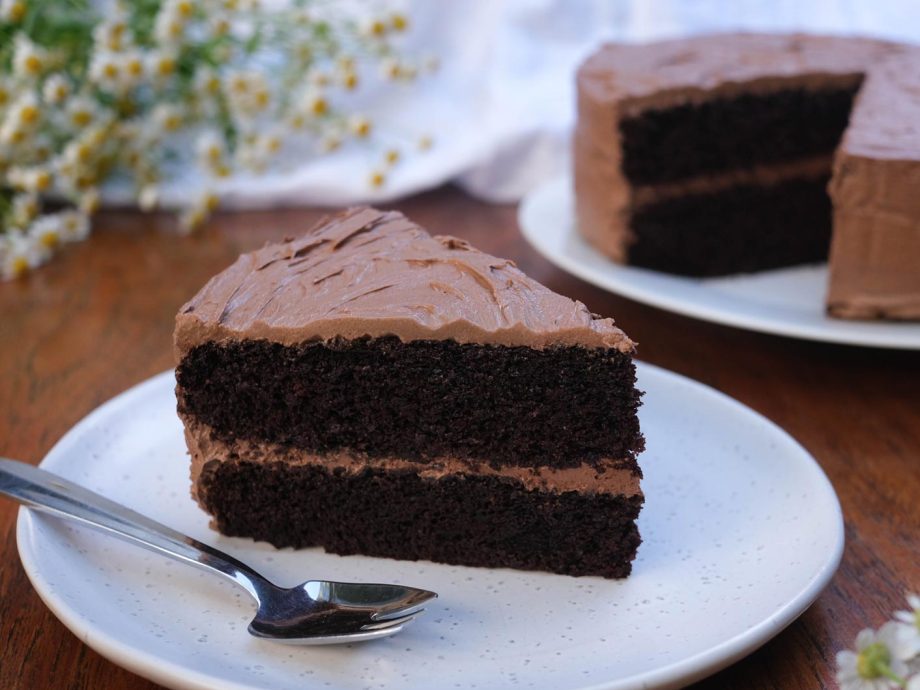 A tall slice of vegan chocolate cake with glossy chocolate frosting on a white plate, crumbs scattered on a warm kitchen counter with soft natural light