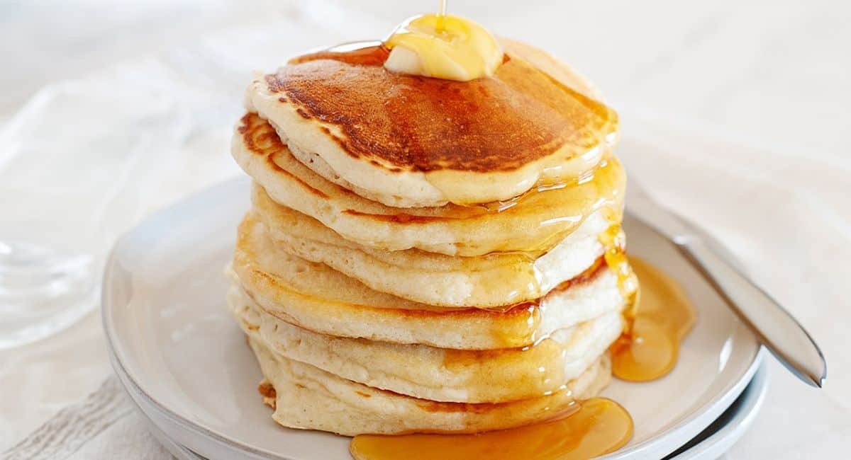 A tall stack of fluffy eggless pancakes on a white plate with butter melting on top and warm maple syrup dripping down, cozy breakfast table setting with soft morning light
