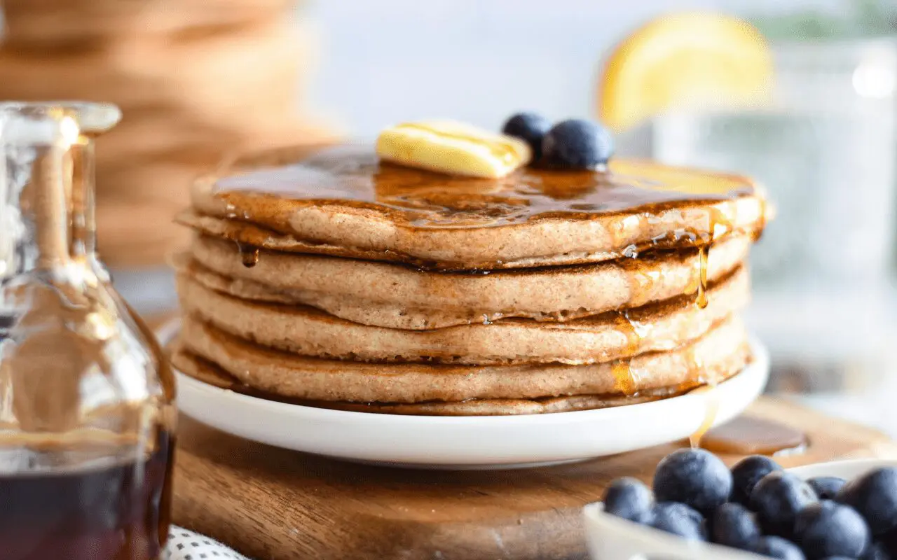 A tall stack of fluffy whole wheat pancakes on a white plate with a pat of butter melting on top and maple syrup pooling around the edges on a sunlit kitchen table
