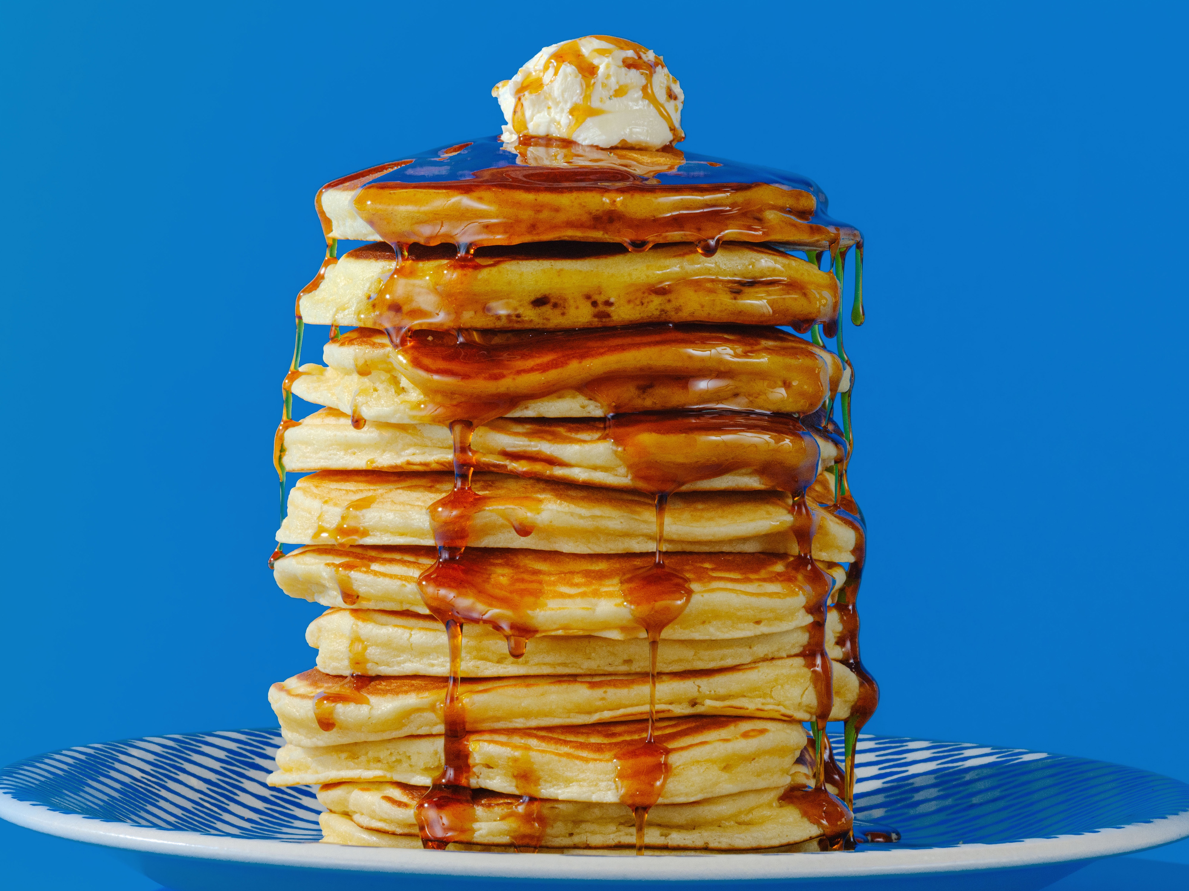 A tall stack of golden buttermilk pancakes on a white plate with butter melting on top and maple syrup drizzling down the sides on a cozy kitchen table