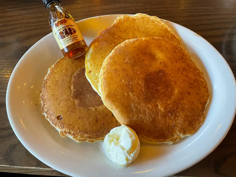 A tall stack of golden buttermilk pancakes on a white plate with a pat of butter melting on top and maple syrup drizzled down the sides on a cozy breakfast table