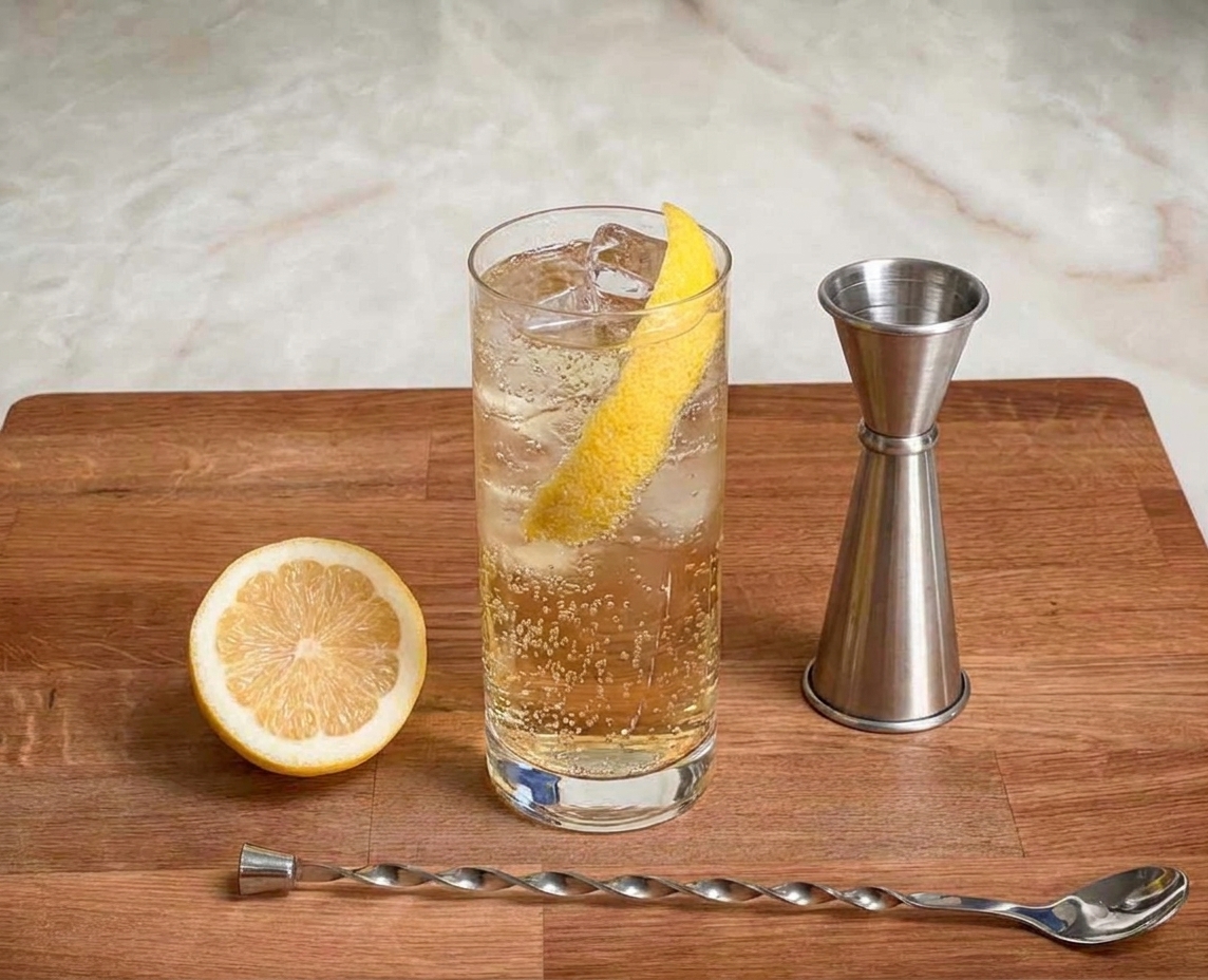 A tall whiskey highball with clear ice, pale golden whiskey, sparkling water bubbles, and a long lemon peel on a kitchen counter