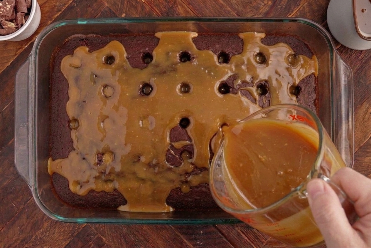 A thick caramel and condensed milk mixture being poured over a poked chocolate cake in a 9x13 pan, with liquid settling into the holes