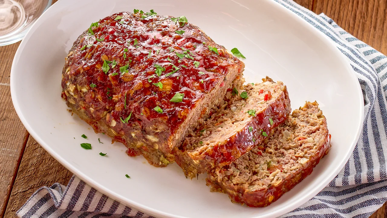A thick slice of Italian meatloaf topped with glossy tomato glaze on a white plate, with a fork and simple side salad in warm kitchen lighting